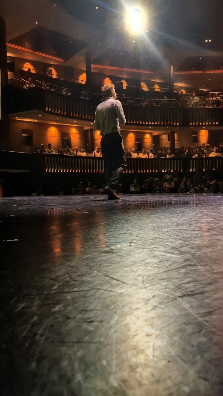 Jacob on stage in a large theatre, facing a full audience across two balconies